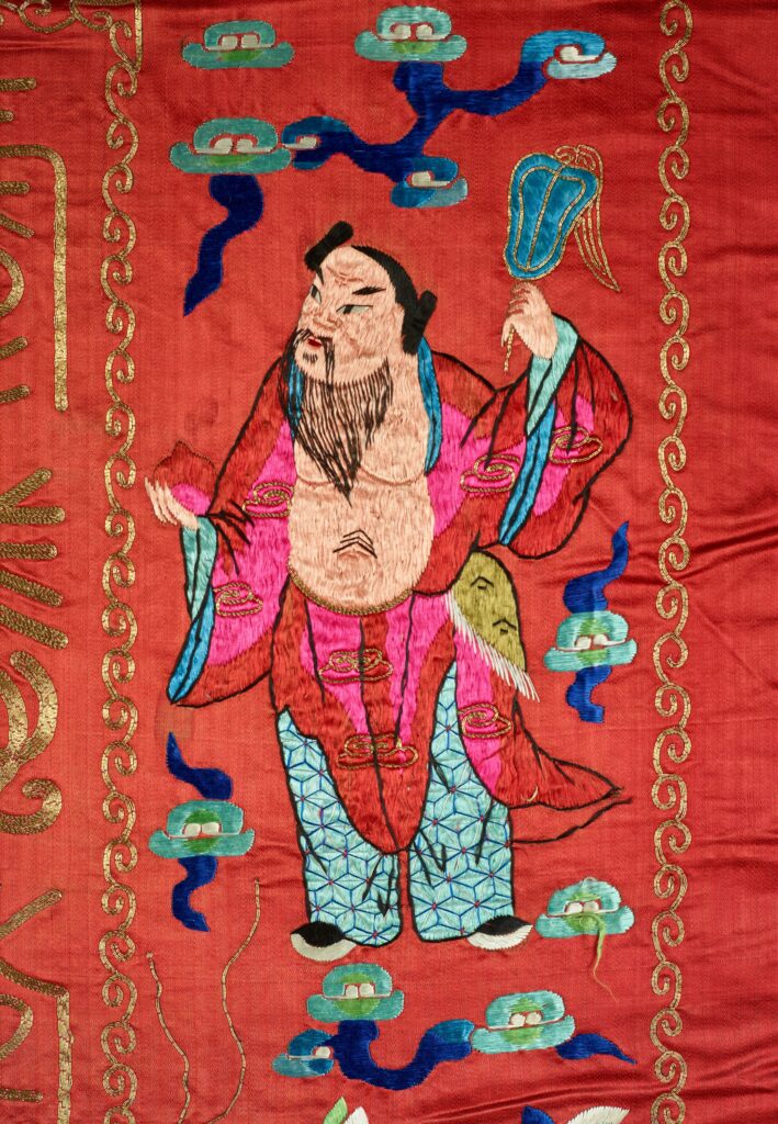 On the tapestry, a bearded male bears his chest and stomach while carrying a blue fan.