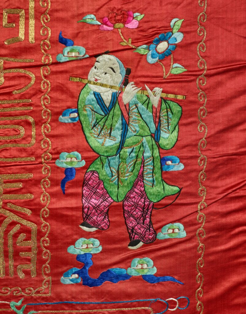 On the tapestry, a young man dressed in green and pink plays a flute with one leg raised.