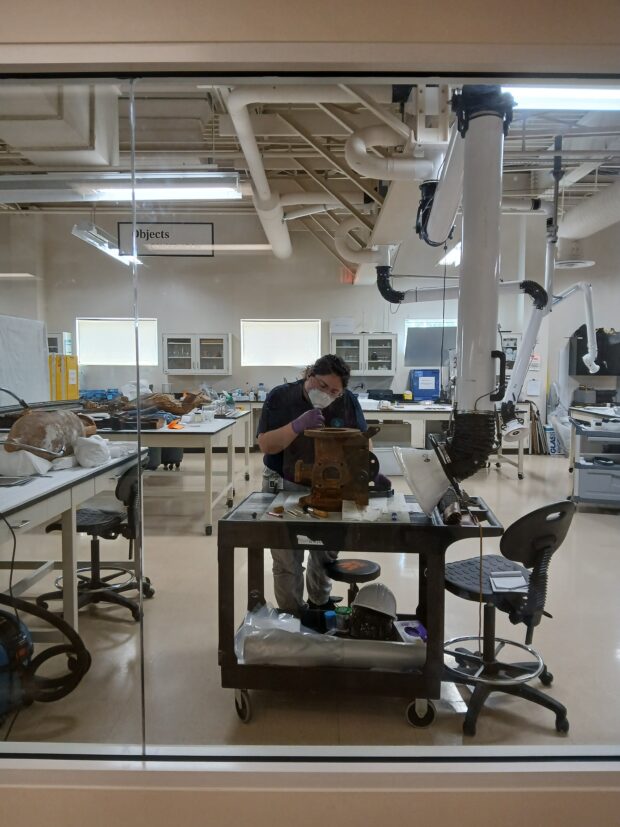 A person in a lab wearing gloves and a mask works on a rusted artifact.