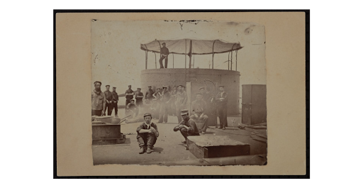 Several men pose for a photo on deck of a ship with one man standing on the turret.