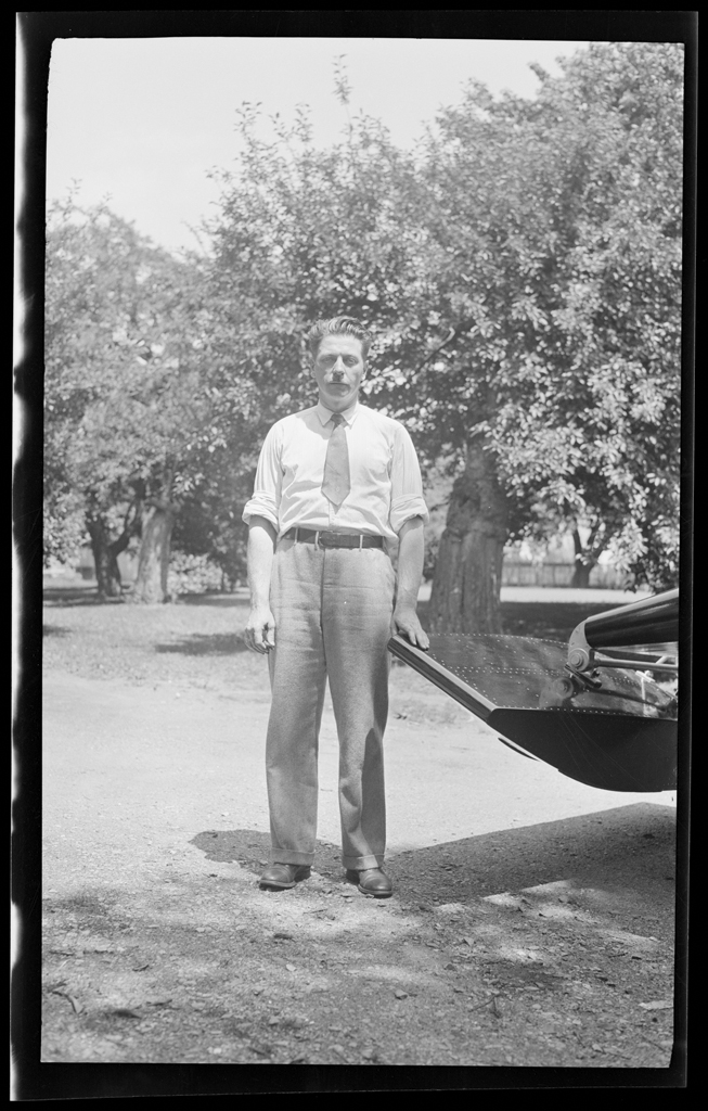 Black and white photo of a man in a tie standing behind a vehicle