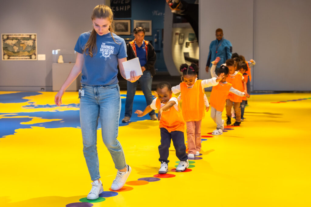 Several children in orange vests walk on circles on the ground, following an adult female in a brightly-colored room