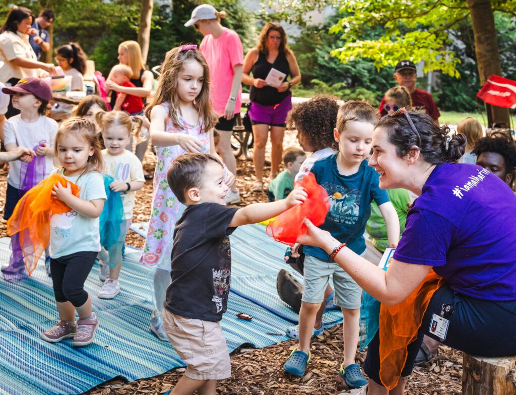 Young children play with colorful scarves on a blanket outdoors and smile at an adult female. Several adults wait in the background.