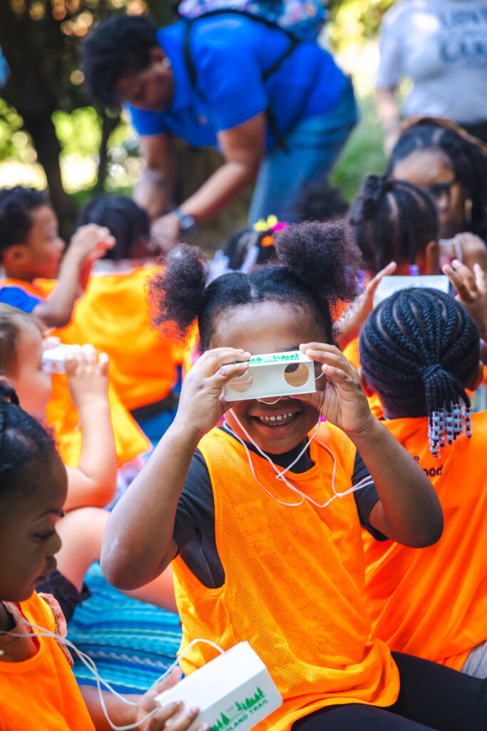 A young girl looks through paper binoculars smiling with other children surrounding her.