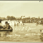 A crowd is in the background watching a group of people dressed in white get baptized in water. Rowboats with men in them float on the same water.