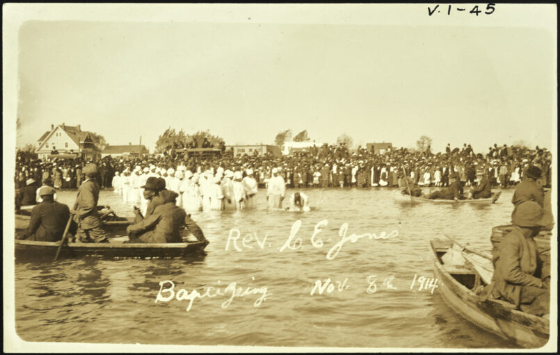 A crowd is in the background watching a group of people dressed in white get baptized in water. Rowboats with men in them float on the same water.