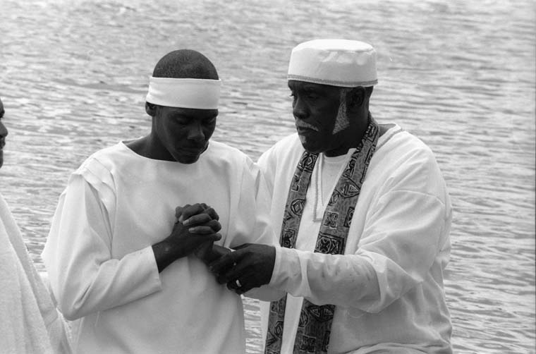 A black a white photo of two men wearing white standing in the water. One man has his hands in the prayer position while the other man touches his arm.